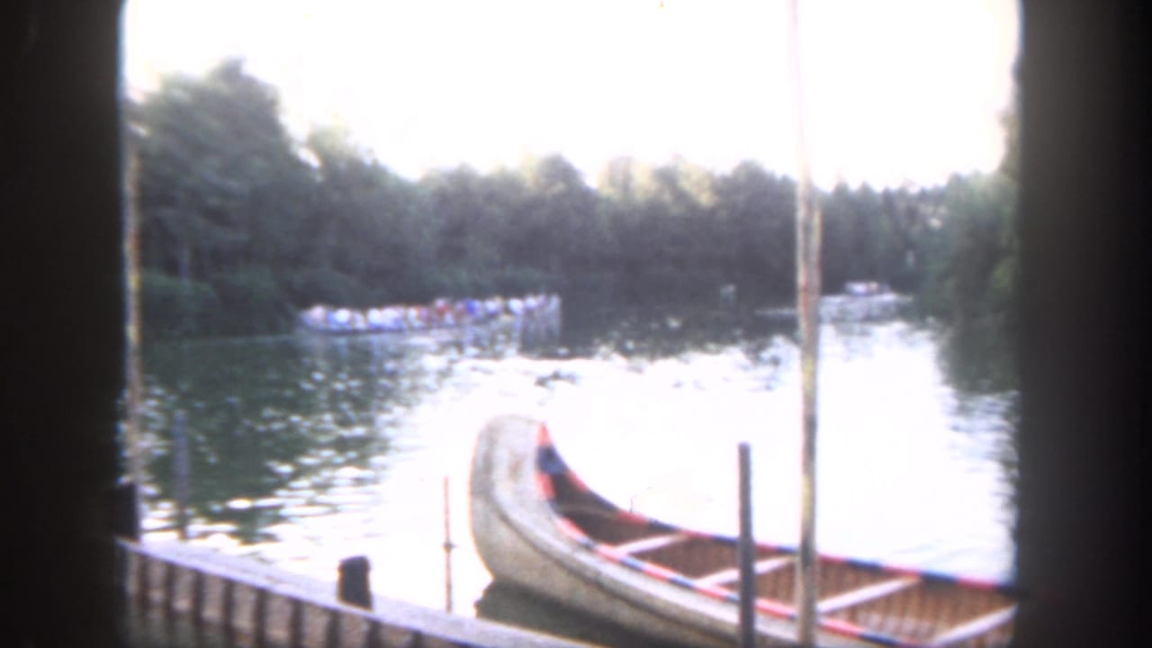 Docked wooden canoe beside water with a distant passenger boat and trees beyond.