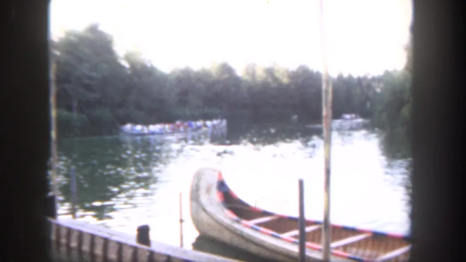 1962 Disneyland home-movie frame showing a wooden canoe at a dock, rippling water, distant guests on a boat, and a wooded shoreline.