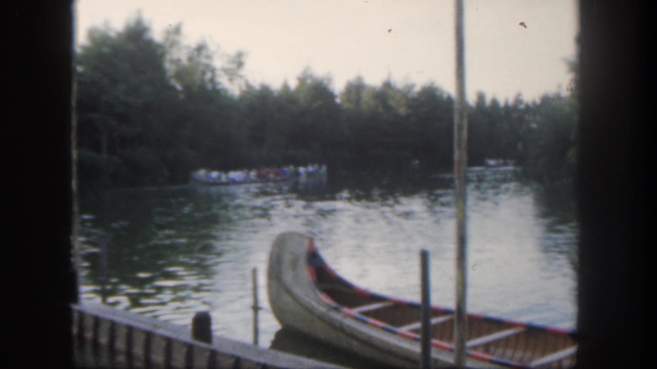Visitor-shot Disneyland waterfront frame with a docked canoe in front and a distant boat of guests near the tree-lined shore.