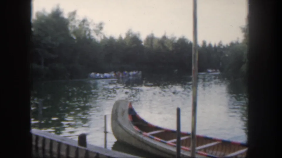 Soft 8mm Disneyland frame showing the canoe lower in view, dock posts, rippling water, and a distant passenger boat near trees.