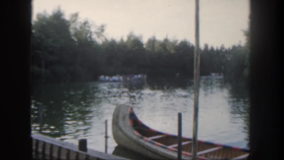 1962 Disneyland home-movie frame with a docked canoe, vertical posts, distant guests on the water, and dark 8mm frame borders.