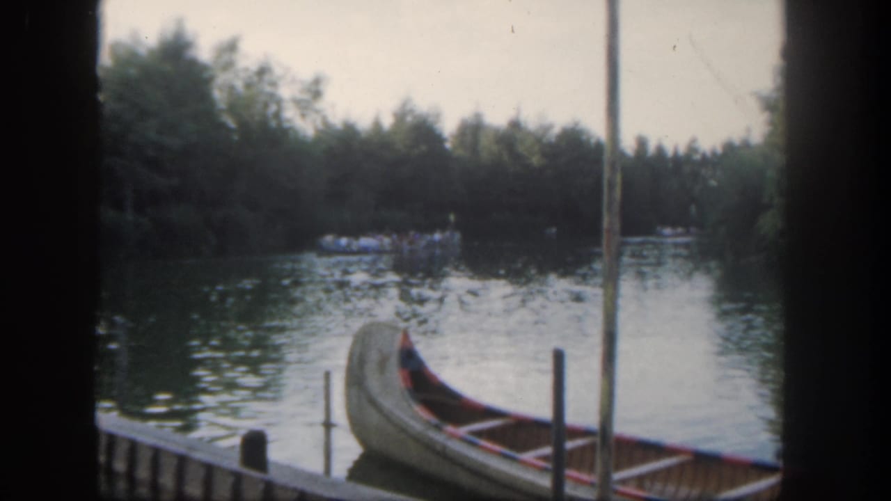 1962 Disneyland home-movie frame showing a canoe at a river dock with wooded shoreline and distant guests on the water.
