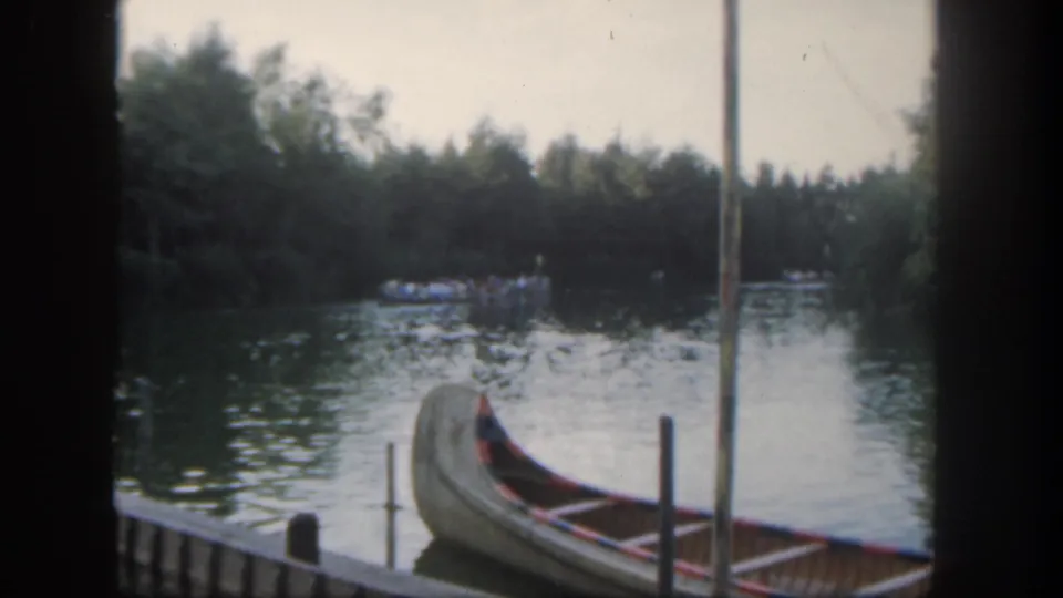 1962 Disneyland home-movie frame showing a canoe at a river dock with wooded shoreline and distant guests on the water.