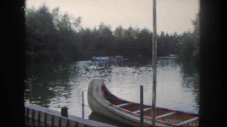 Visitor-shot 1962 Disneyland footage frame showing a canoe by the dock and a guest boat farther along the wooded river.