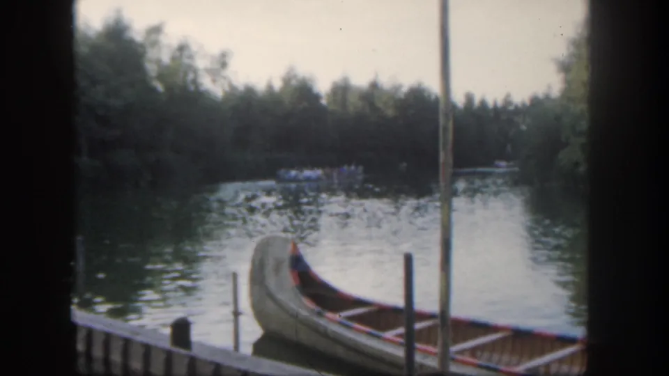 1962 Disneyland home-movie frame with a docked canoe, vertical posts, reflective water, and a distant boat near the tree line.