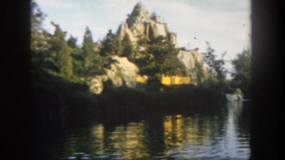1962 Disneyland boat-ride view across calm water toward artificial rockwork, trees, and reflected scenery.