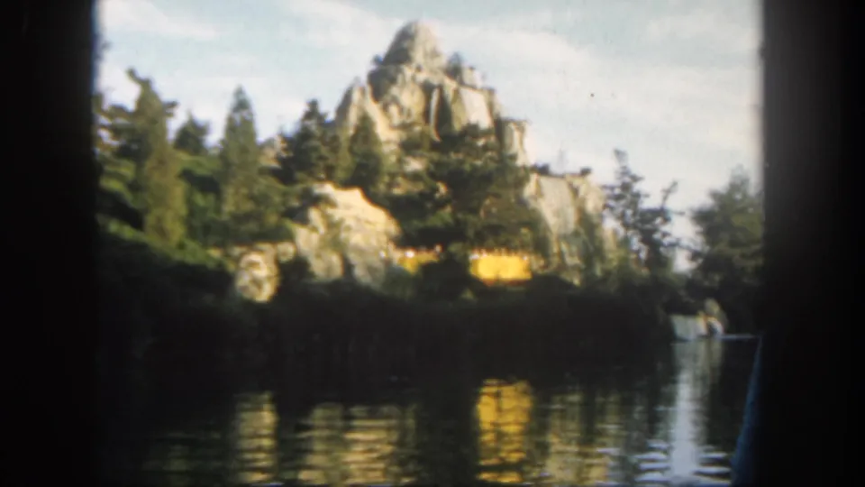 Disneyland 1962 home-movie frame showing artificial rockwork, dense trees, calm water, and a small cascade-like feature.