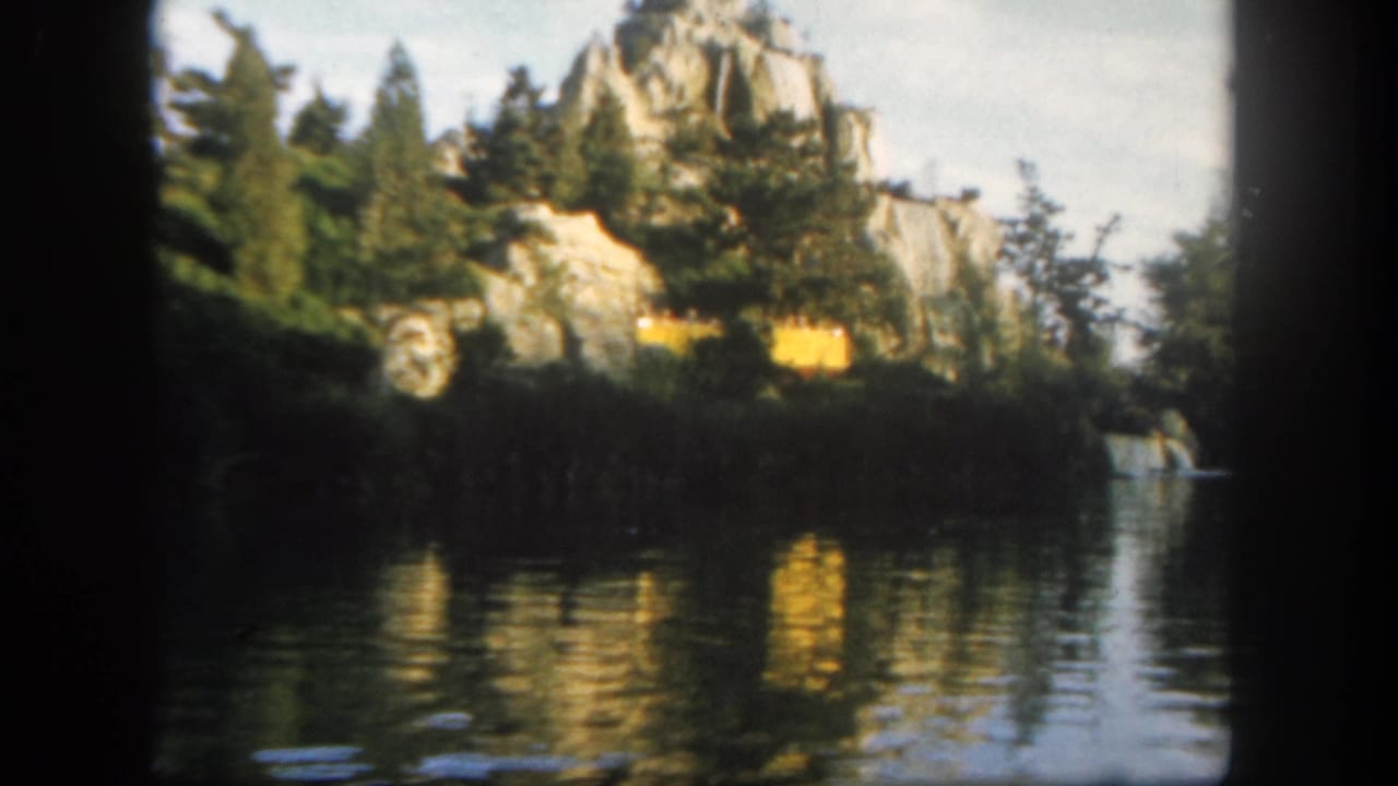 1962 Disneyland home-movie frame showing a canoe on calm water below rocky scenery and evergreen planting.