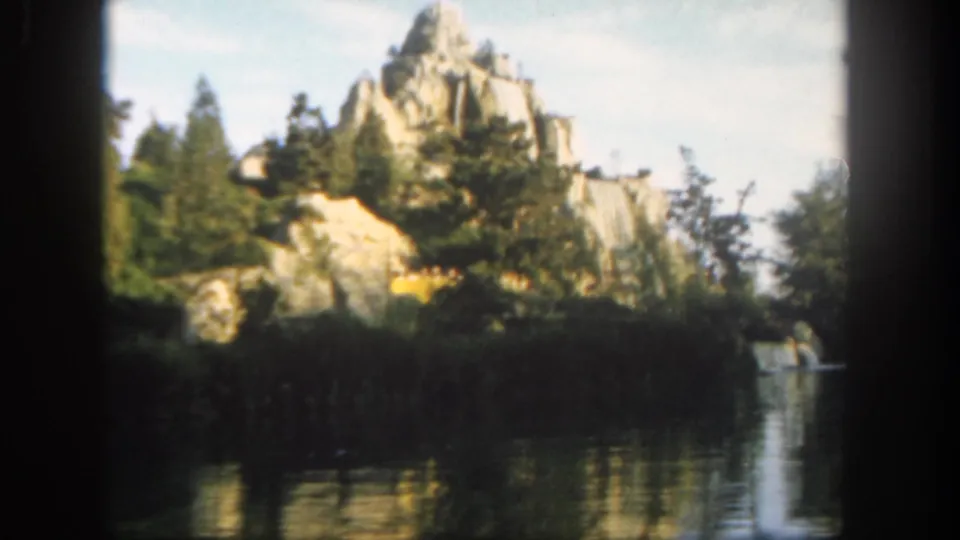1962 Disneyland 8mm frame showing bright rocky scenery, evergreens, reflective water, and a canoe near the right side.