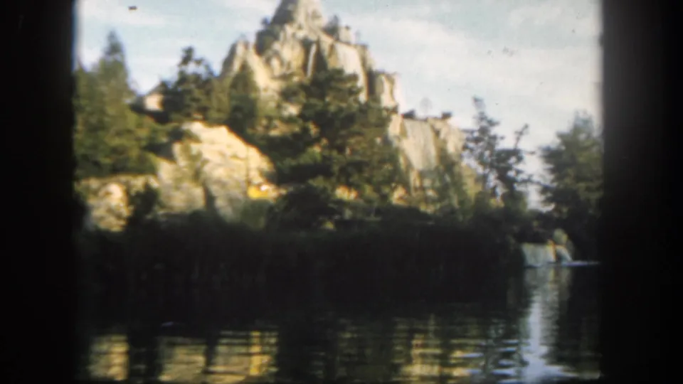 1962 Disneyland home-movie still with a canoe by the riverbank, rocky backdrop, evergreen trees, and soft water reflections.
