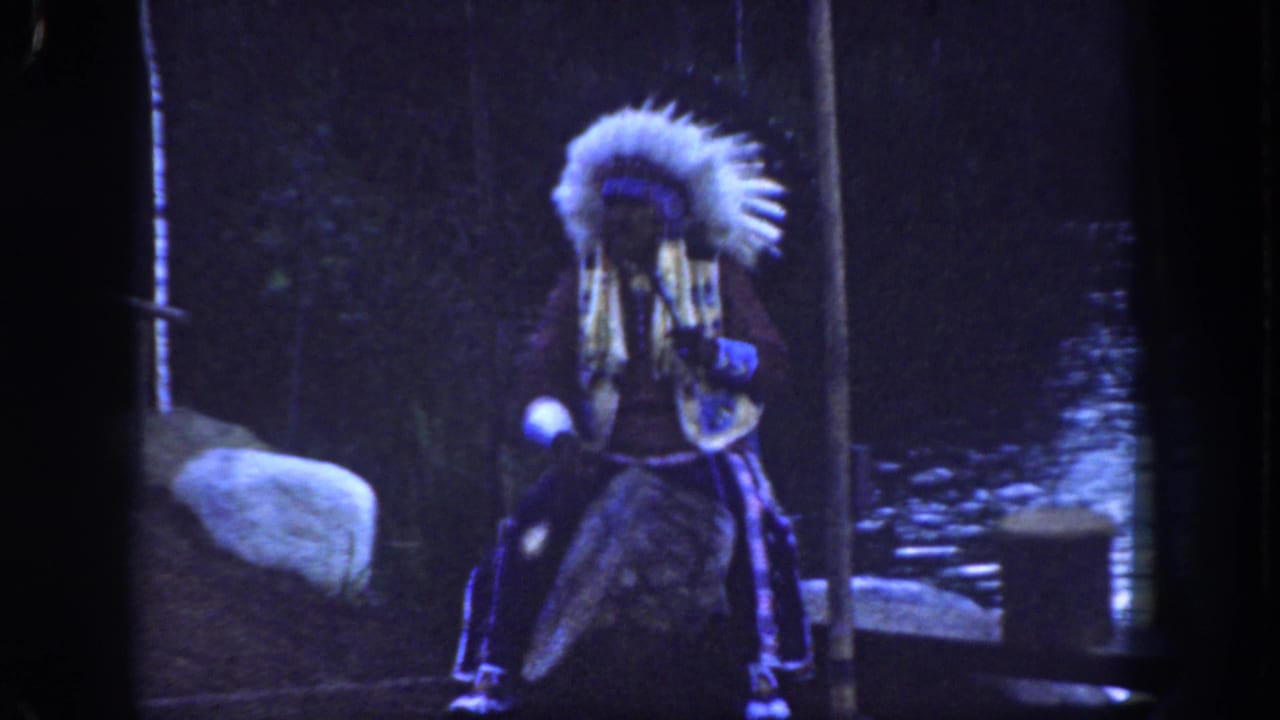 Performer in feathered headdress stands with a flute-like instrument near rocks and water.