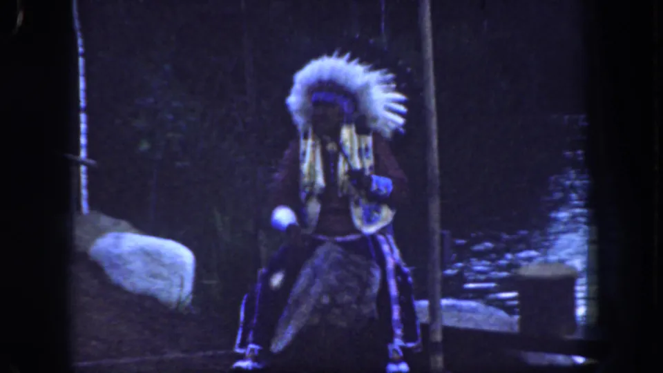 1962 Disneyland home-movie frame showing a Native American performer in regalia holding a flute-like instrument in a wooded outdoor set.