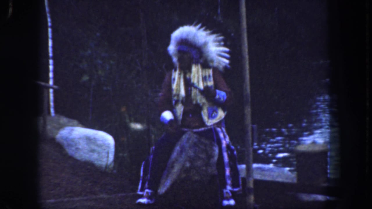 Performer faces partly sideways in feathered regalia beside a rock and dark water.