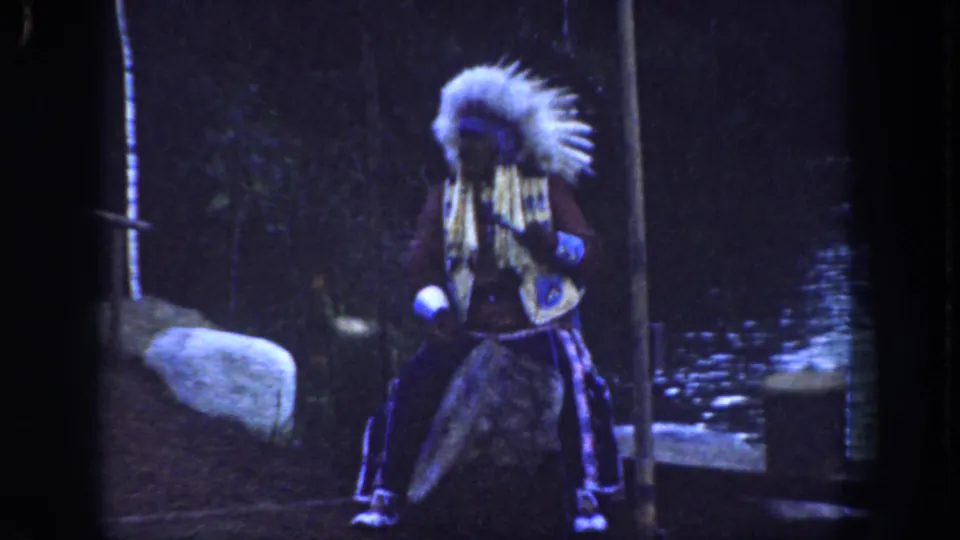 1962 Disneyland frame with a performer in feathered regalia turned sideways, holding a flute-like instrument in a shaded wooded set.
