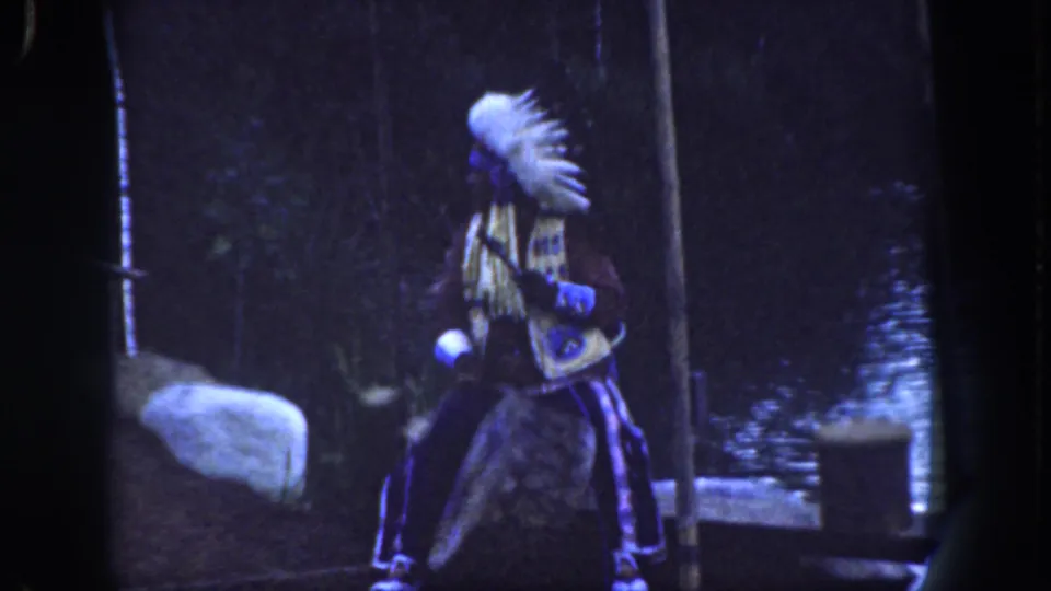 Visitor-shot 1962 Disneyland frame showing a regalia-clad performer on a small platform in a wooded set with water and rock scenery.