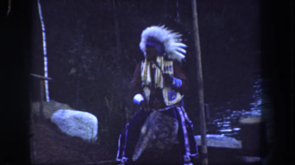 1962 Disneyland 8mm frame of a Native American performer in feathered regalia holding a flute-like instrument near water and wooded scenery.