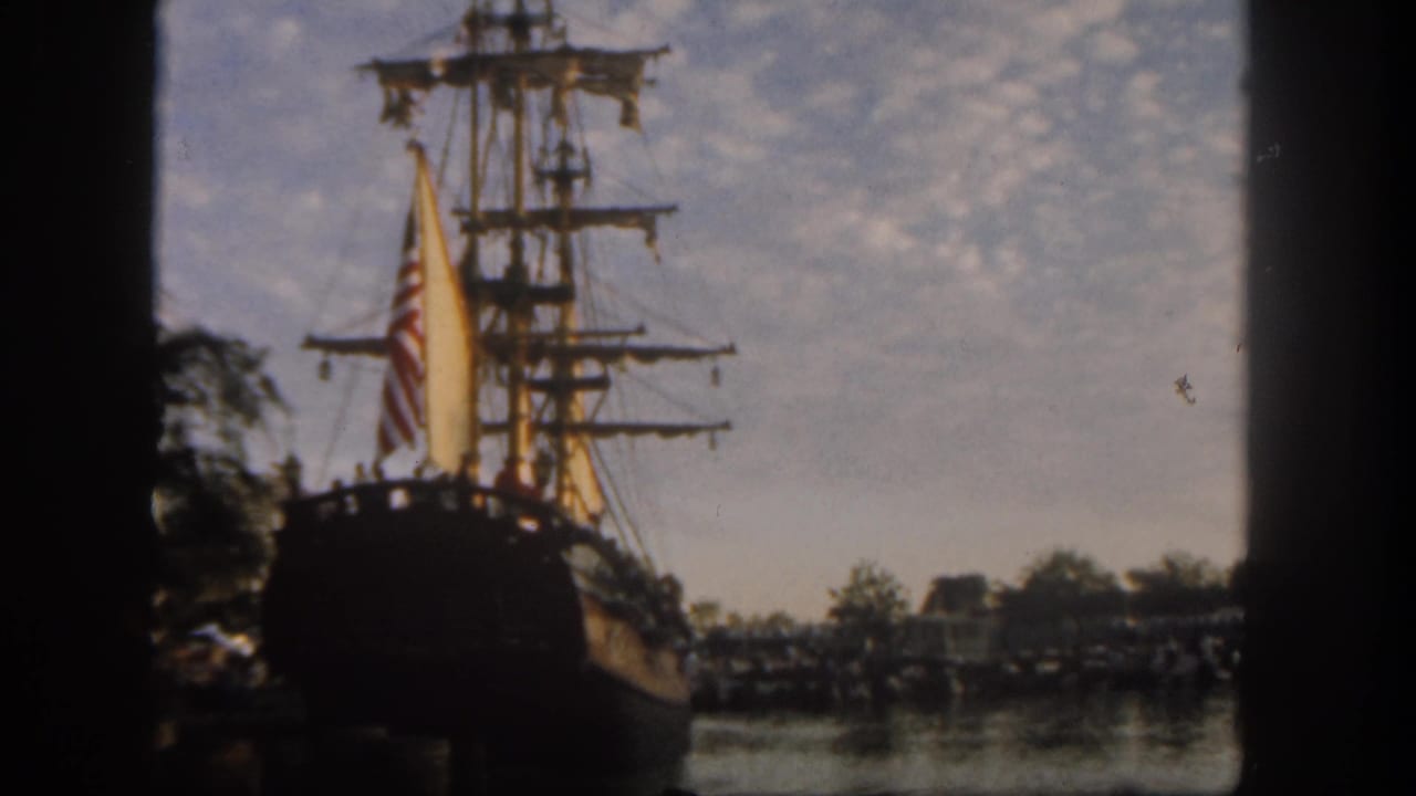 1962 Disneyland home-movie frame showing a docked sailing ship with pale sails and an American flag beside a reflective waterfront.
