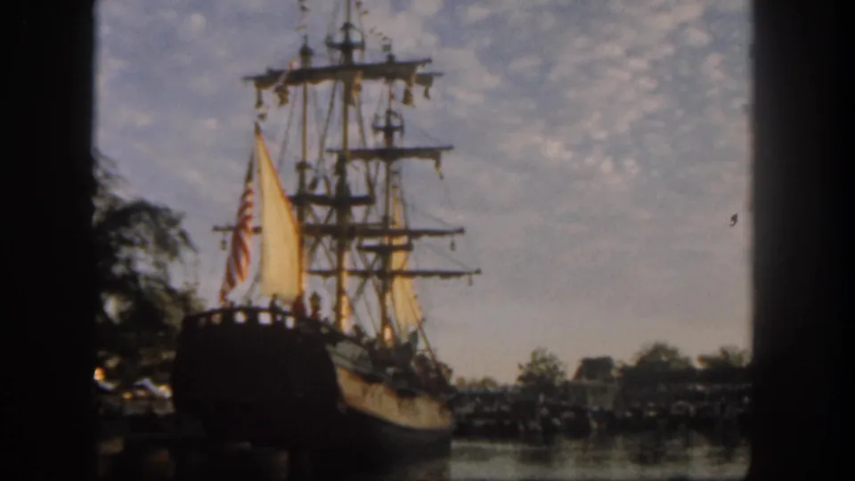 1962 Disneyland waterfront frame with a tall wooden sailing ship, flag, trees, and water reflections under a mottled sky.