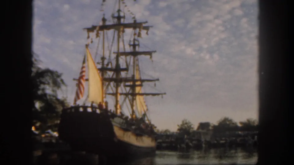 1962 Disneyland home-movie frame showing a tall-masted wooden sailing ship with yellow sails, American flag, water, trees, and distant visitors.