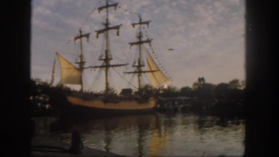 1962 Disneyland home-movie frame showing the sailing ship in side view, with masts, yellow sails, shoreline crowds, water reflections, and cloudy sky.