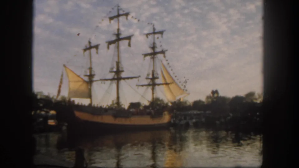 1962 Disneyland frame showing the wooden sailing ship from the side, with rigging, yellow sails, calm water, shoreline visitors, trees, and open sky.