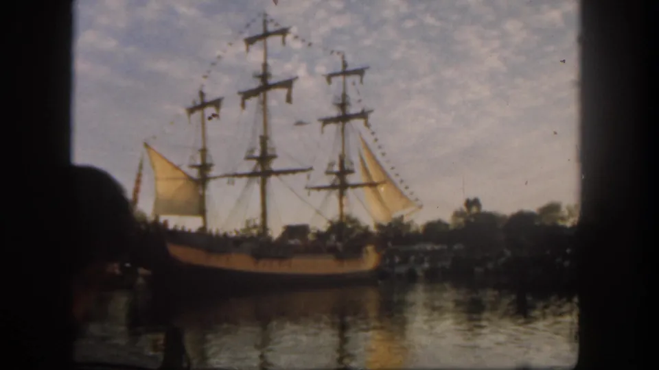 1962 Disneyland home-movie frame showing a sailing ship with pale sails moving across reflective water.