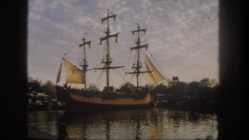 1962 Disneyland footage still showing a multi-masted ship moving across water with distant visitor silhouettes nearby.