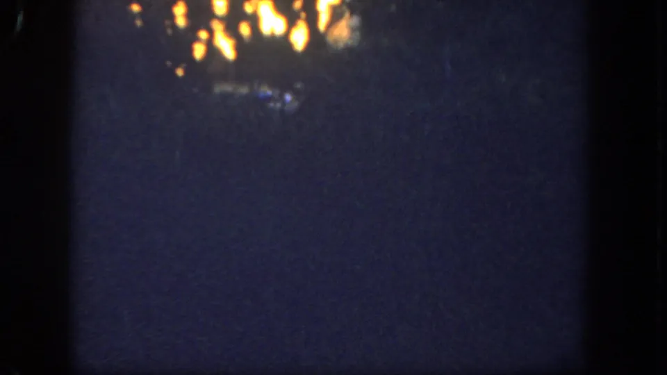 Underexposed 8mm frame from the 1962 Disneyland clip, with partial firelight visible above a dark wooded foreground.