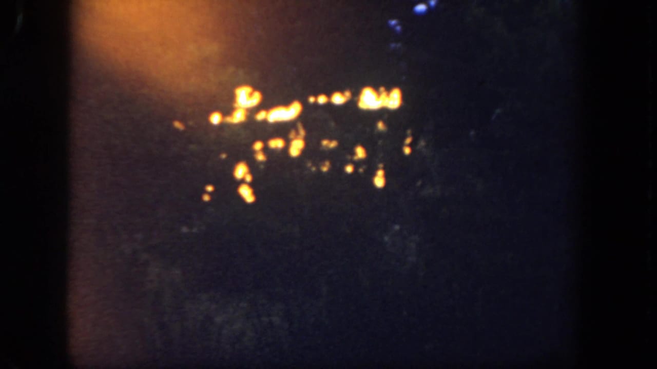 Dim 1962 Disneyland night frame showing warm glowing lights with a soft orange exposure flare.
