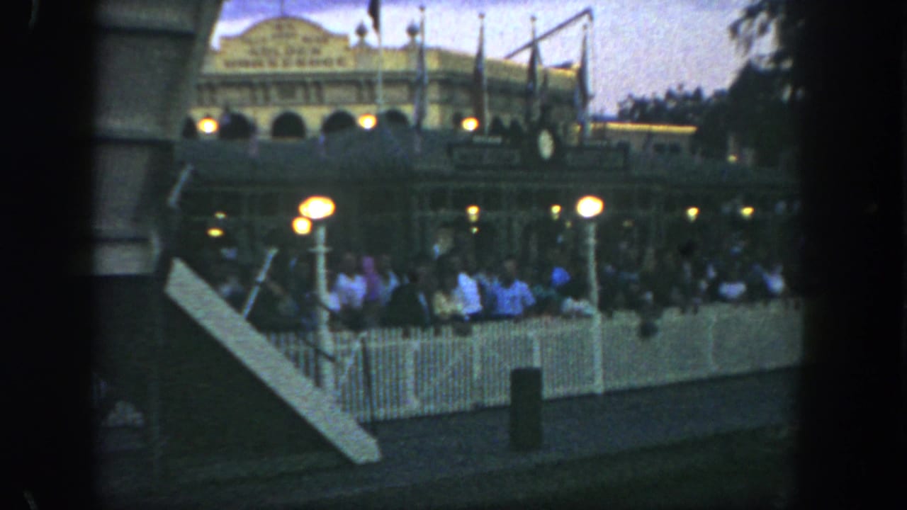 1962 Disneyland visitors wait behind white picket fencing near warm globe lamps with the Golden Horseshoe facade visible beyond.