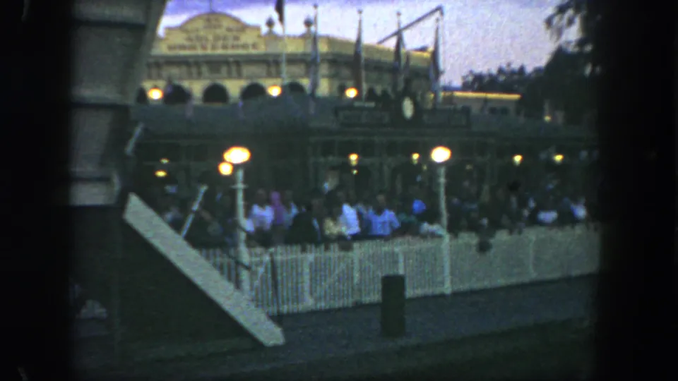 1962 Disneyland visitors wait behind white picket fencing near warm globe lamps with the Golden Horseshoe facade visible beyond.