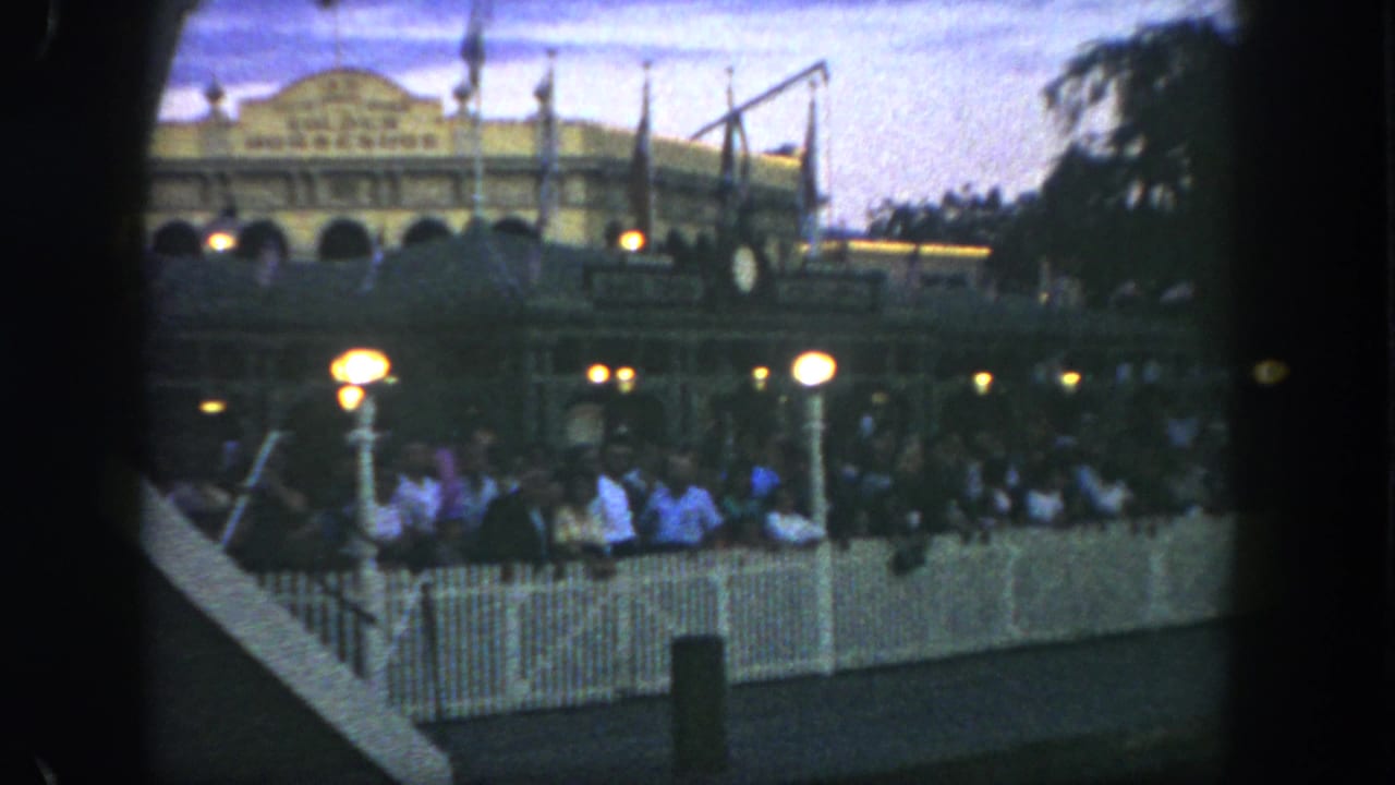 Disneyland guests stand in a fenced queue at dusk, with lit globe lamps and the Golden Horseshoe building visible behind them.