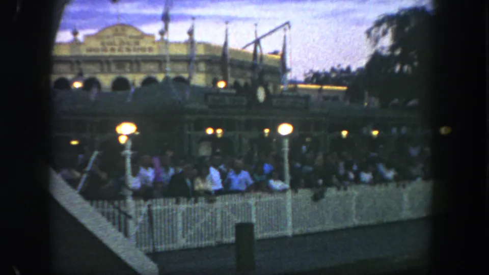 Disneyland guests stand in a fenced queue at dusk, with lit globe lamps and the Golden Horseshoe building visible behind them.