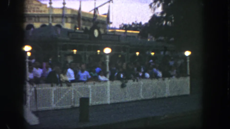 The 8mm frame shows a Disneyland queue area with guests behind white fencing, glowing lamps, stairs, and the Golden Horseshoe facade.