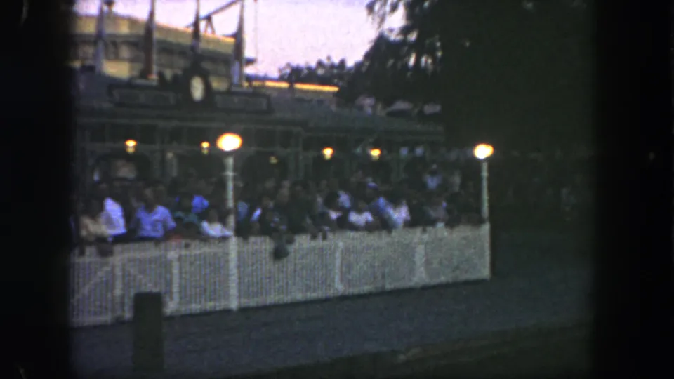 A dimmer 1962 Disneyland view shows guests waiting behind white fencing, with bright lamps and the Golden Horseshoe area partly obscured by trees.