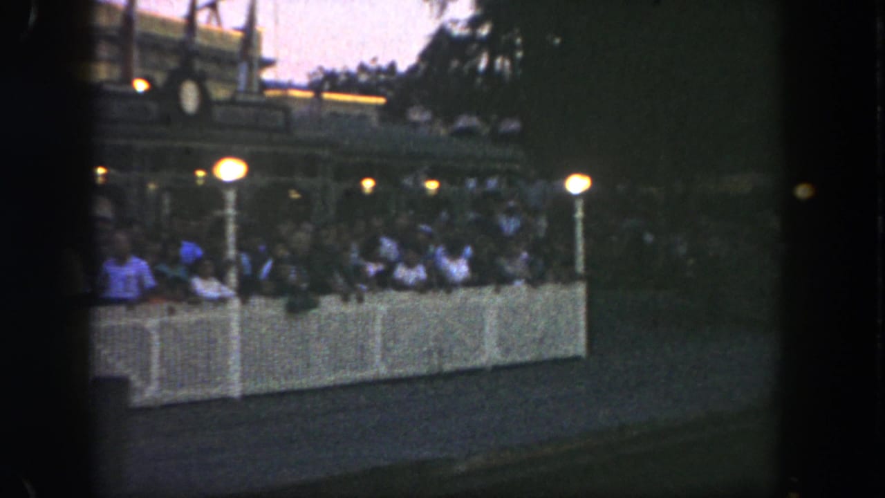 Nighttime crowd behind a white barrier near lamp posts and a paved walkway.