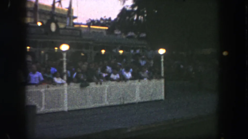 1962 Disneyland home-movie frame showing a crowd behind a white barrier under warm park lamps at night.