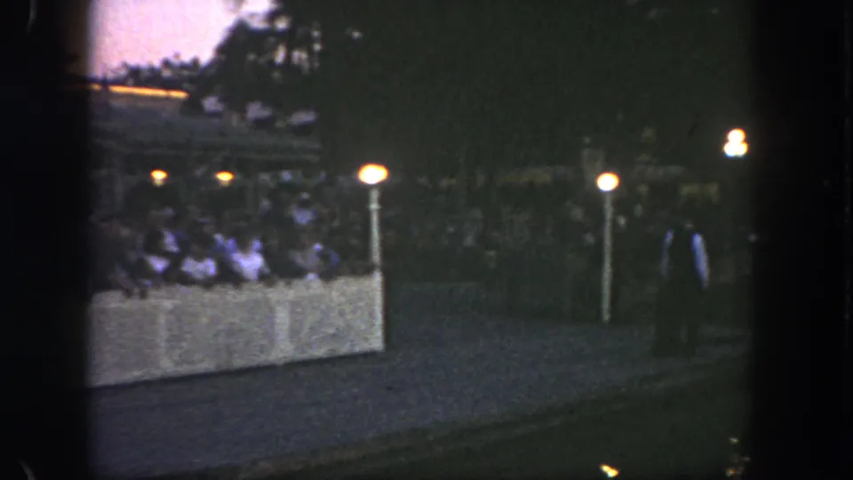 Nighttime 8mm view at Disneyland with visitors behind a barrier, lit posts, and a figure near the walkway.