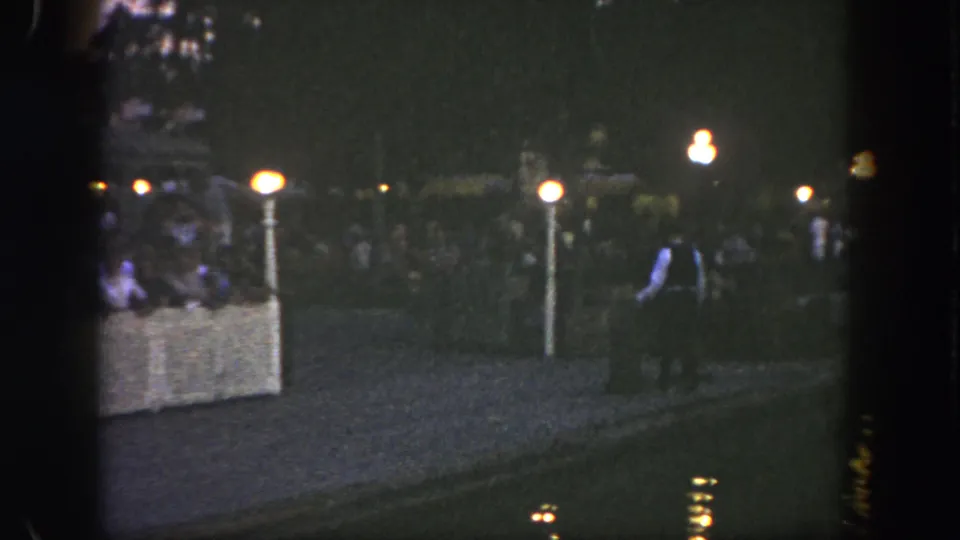 1962 Disneyland nighttime frame with a person in a light shirt walking past lamps and a distant crowd.