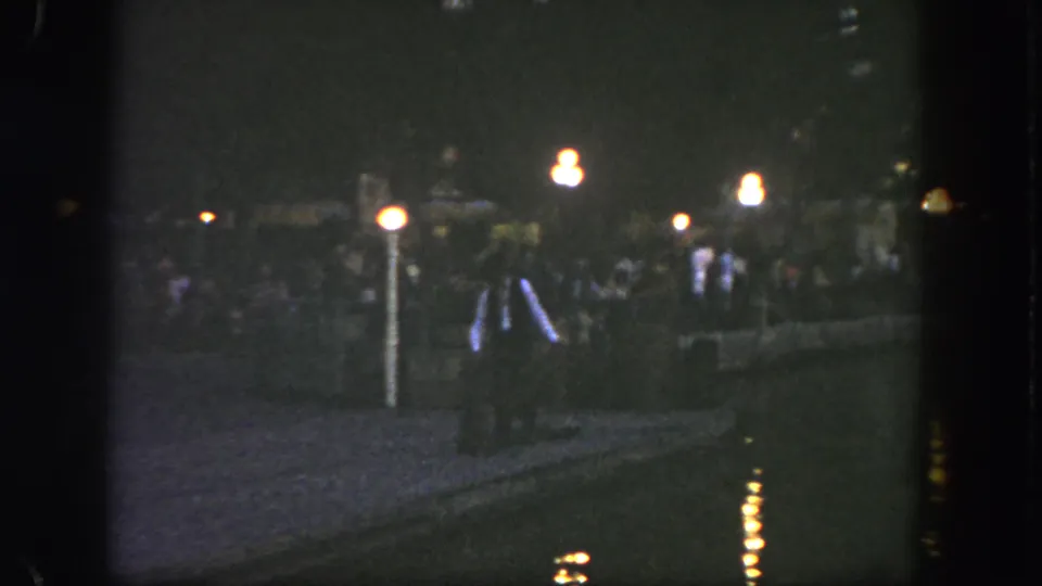 Low-light Disneyland home-movie frame showing a walker near the path, lamp glow, reflections, and distant visitors.