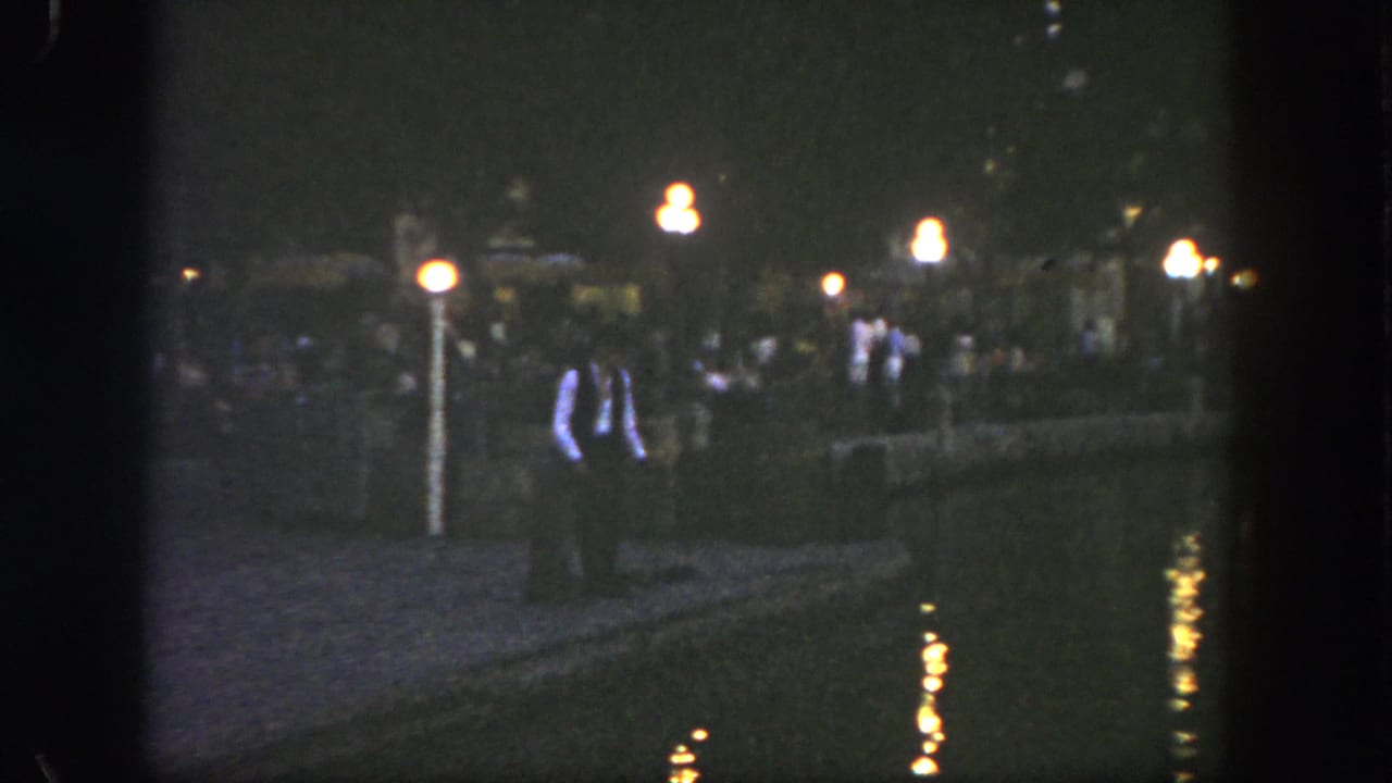 1962 Disneyland home-movie frame showing visitors along a dark riverside walkway with warm lamp reflections on the water.