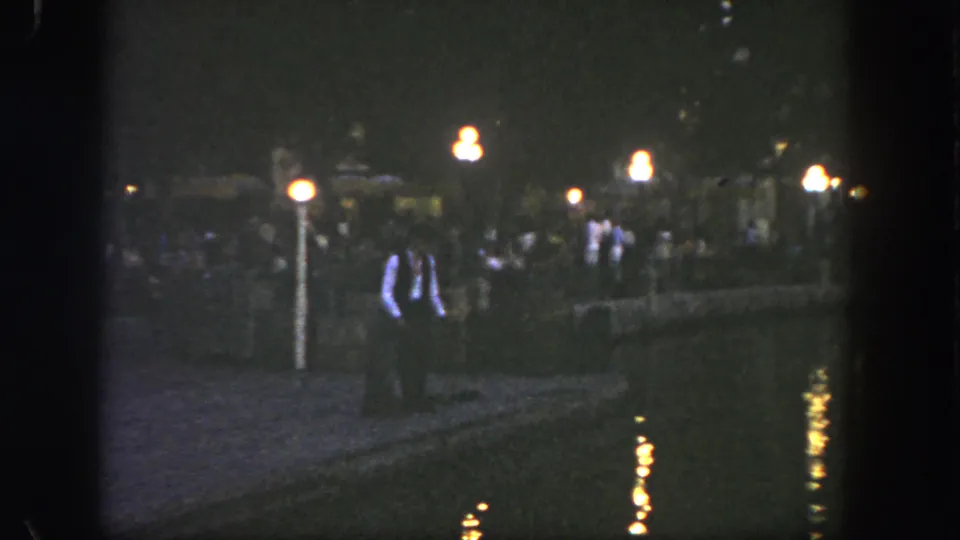 1962 Disneyland home-movie frame showing visitors along a dark riverside walkway with warm lamp reflections on the water.