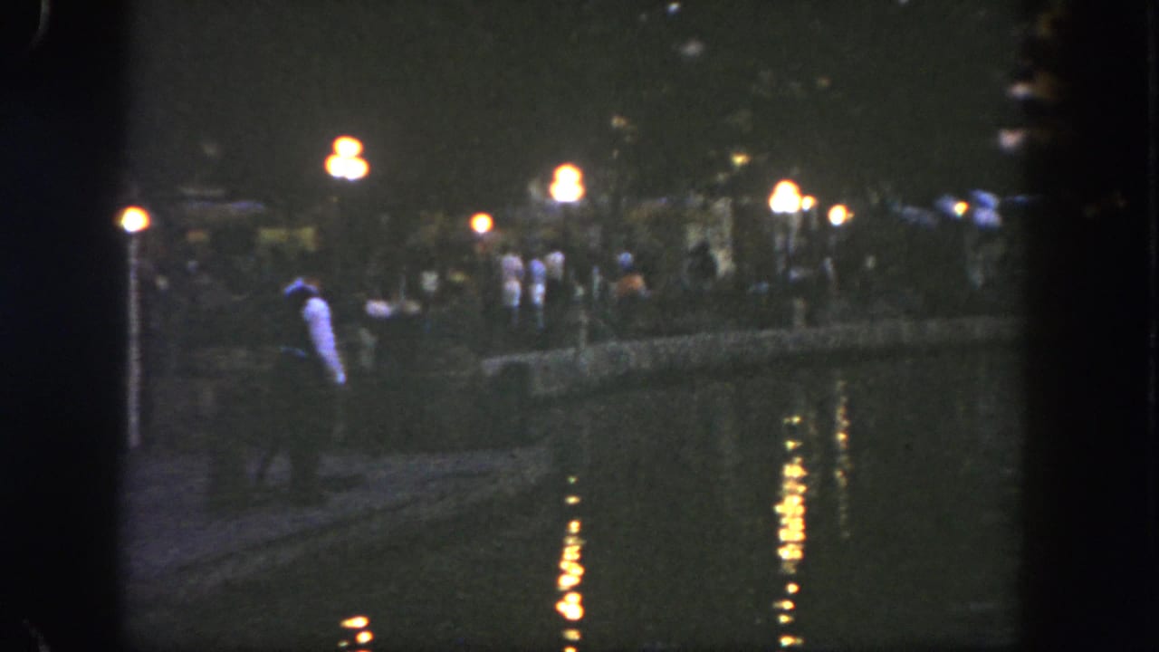 1962 Disneyland frame with a light-shirted man near the waterfront and visitors gathered along the lamp-lit path.