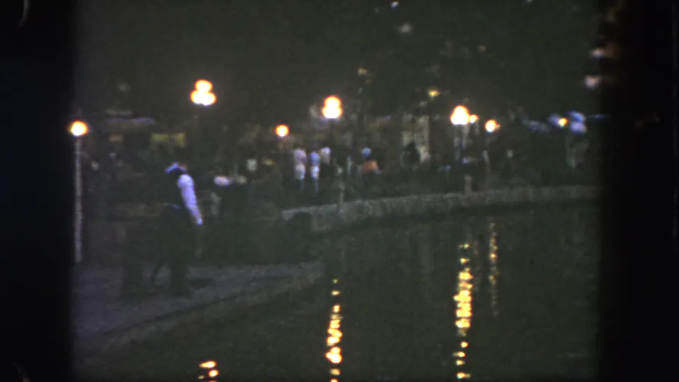 1962 Disneyland frame with a light-shirted man near the waterfront and visitors gathered along the lamp-lit path.