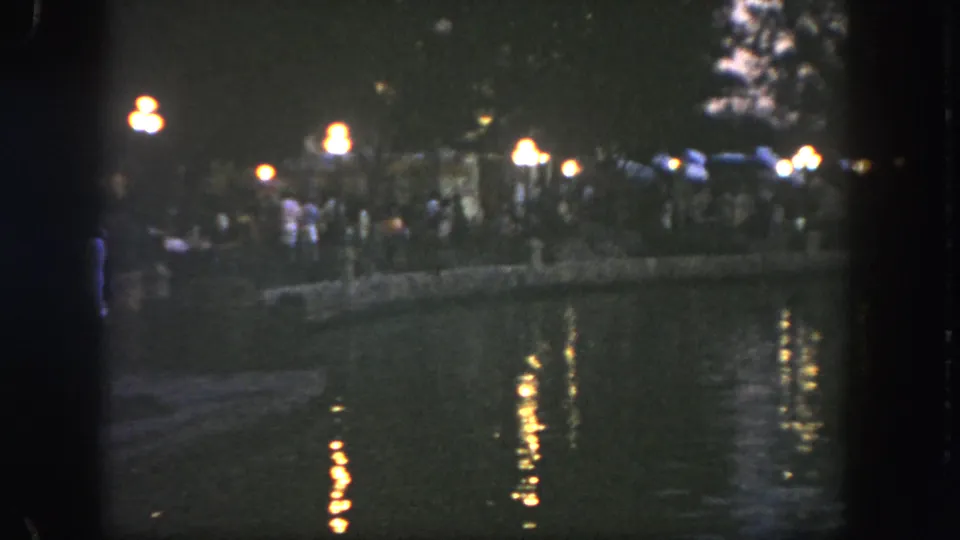 1962 Disneyland home-movie frame showing a busy waterside promenade with lamps, trees, and reflections in low evening light.