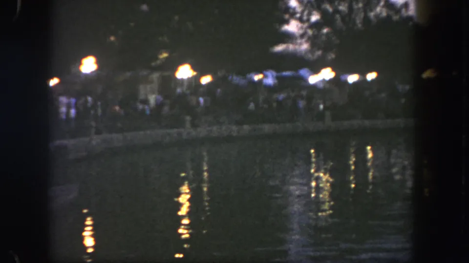 1962 Disneyland frame showing water reflections, visitors on a riverside path, and warm lamps in a dark evening setting.