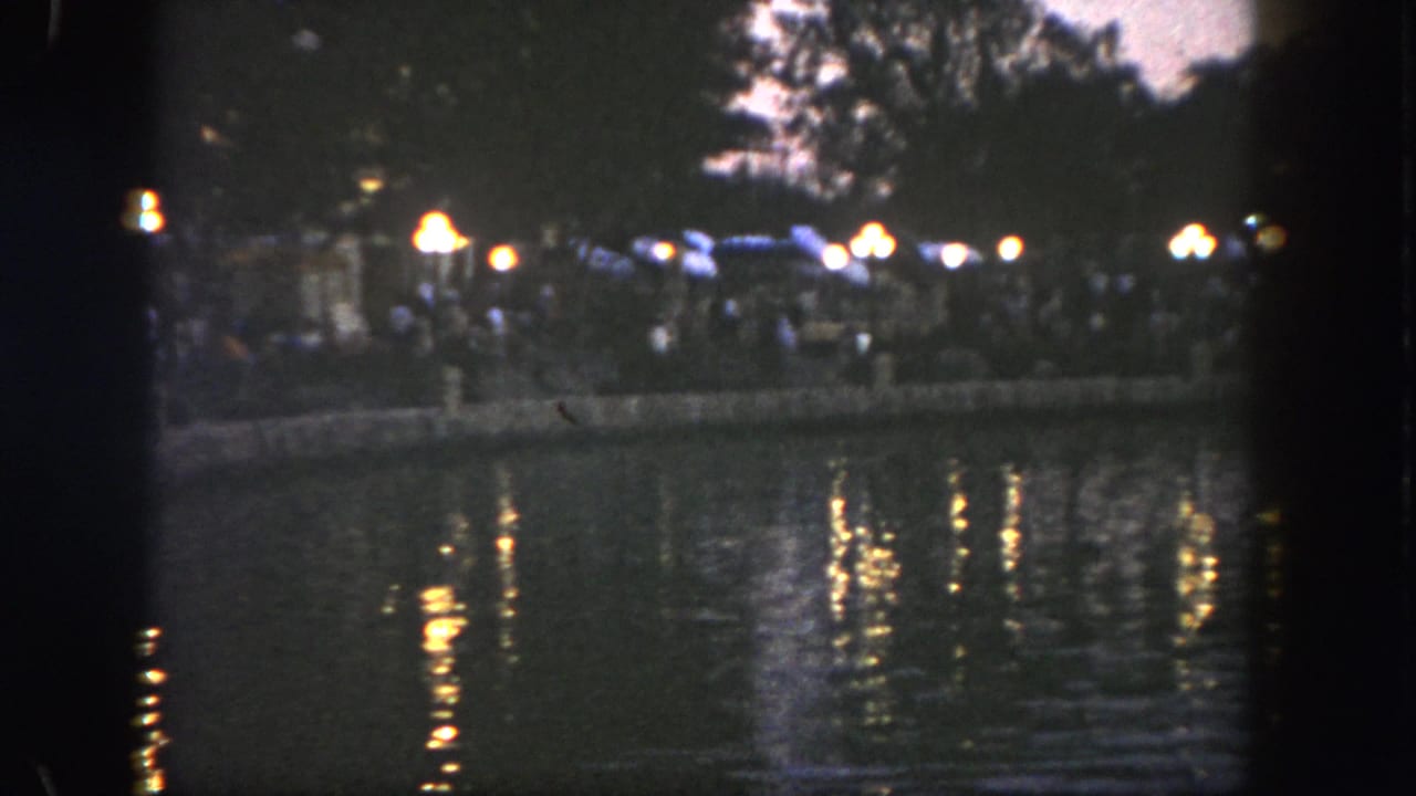 1962 Disneyland home-movie frame showing distant shoreline lights reflected across a dark lake at night.
