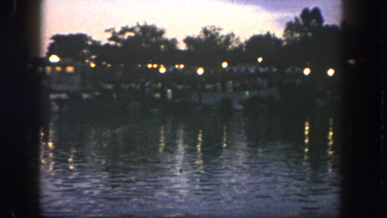 Dusk waterfront with lights reflecting on rippling water and dark trees along the far shore.
