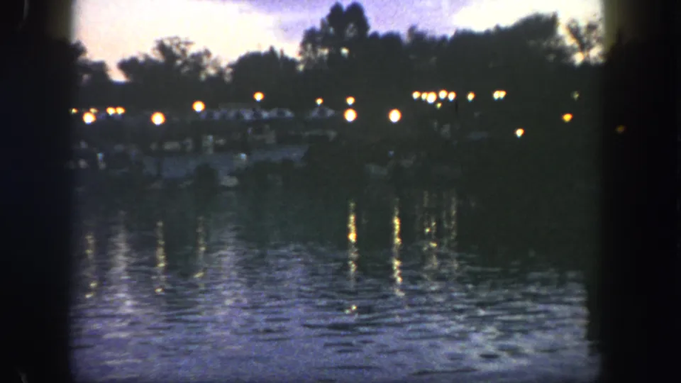 Visitor-shot 1962 Disneyland bay scene with glowing shoreline lights, dark trees, and softly exposed reflections on rippling water.