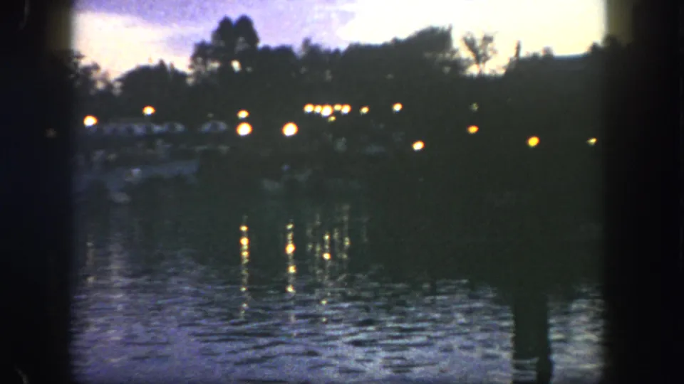 1962 Disneyland dusk waterfront frame with a dark shoreline, clustered lights, and reflected glow across the bay.